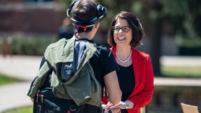 Chancellor Gallo, a woman wearing a red blazer, black top, string of pearls and black glasses smiles and greets a student who has their back to the camera. The student is wearing headphones and has a backpack and jacket on their back. 