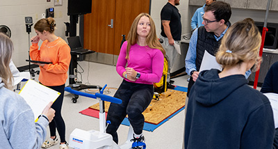 A group of students stand around another student sitting on an exercise bike