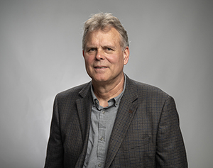 Dave Bonko Headshot. White man with slightly greying hair poses for a head shot wearing a dark grey sport coat with a grey button up shirt in front of a neutral backdrop.