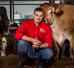 Kurt Vogel headshot. Kurt kneels inside a cow barn and a cow is behind him