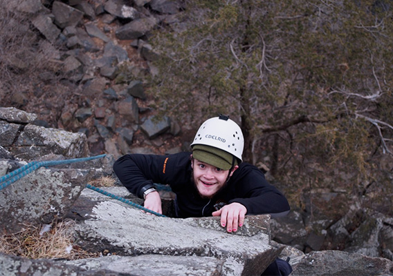 A male student uses climbing equipment to climb a small cliff and smiles up at the camera