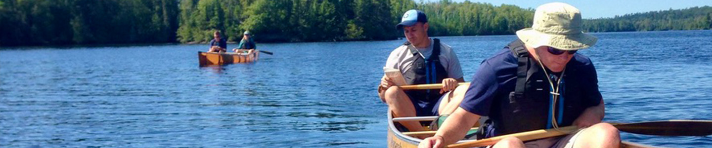 Two male students sit in a canoe and two other students are behind them in the background