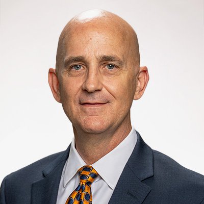 Robert Jaynes headshot. A white man with a bald head smiles directly at the camera in front of a grey background. He is wearing a navy blue suit jacket, white collared shirt and an orange and navy checkerboard tie.